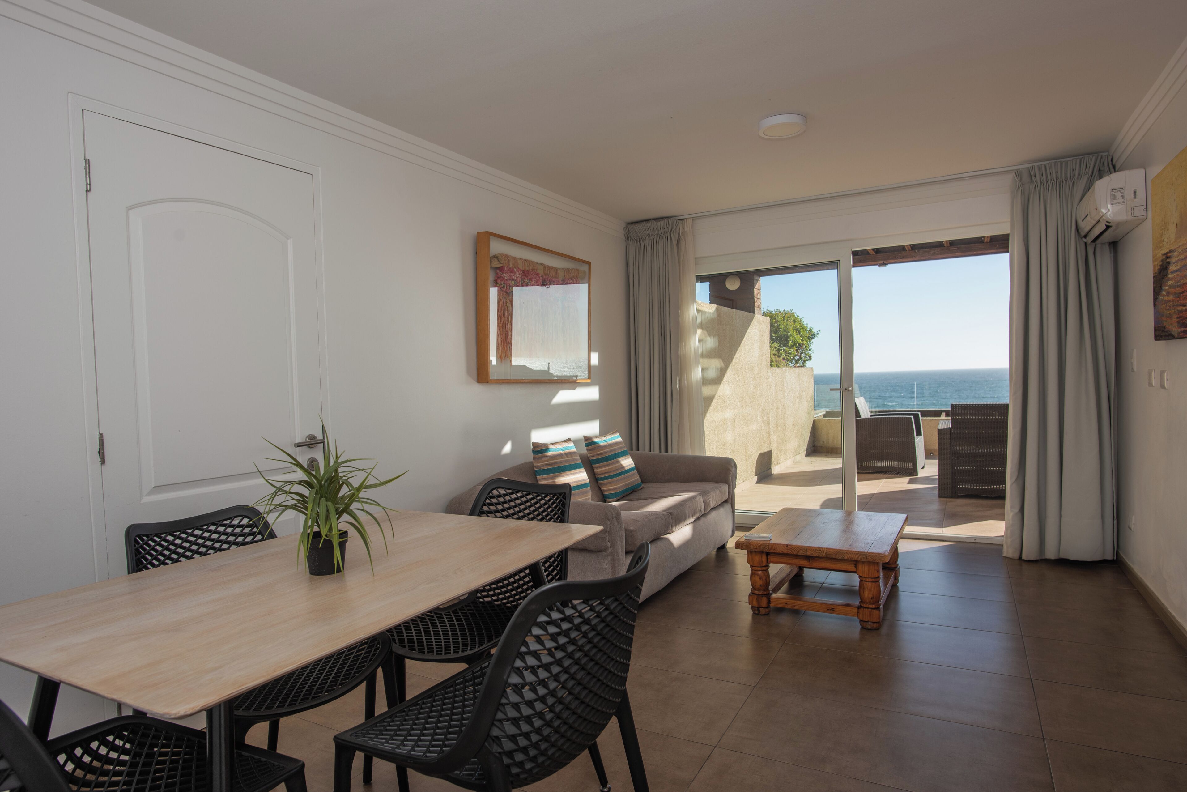 A room with a dining table, chairs, a sofa, a small table, and a view of the sea through a sliding door.