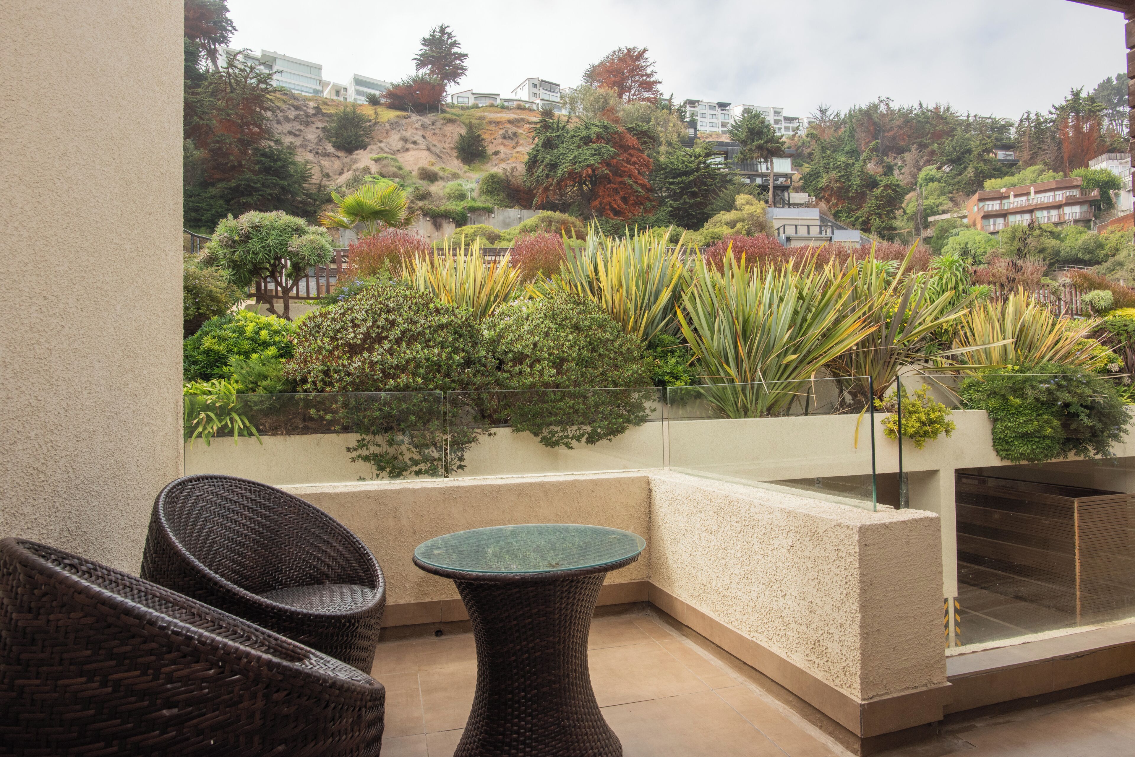 A balcony with wicker furniture, a glass table, and a view of a landscaped garden with various plants and a hillside in the background.