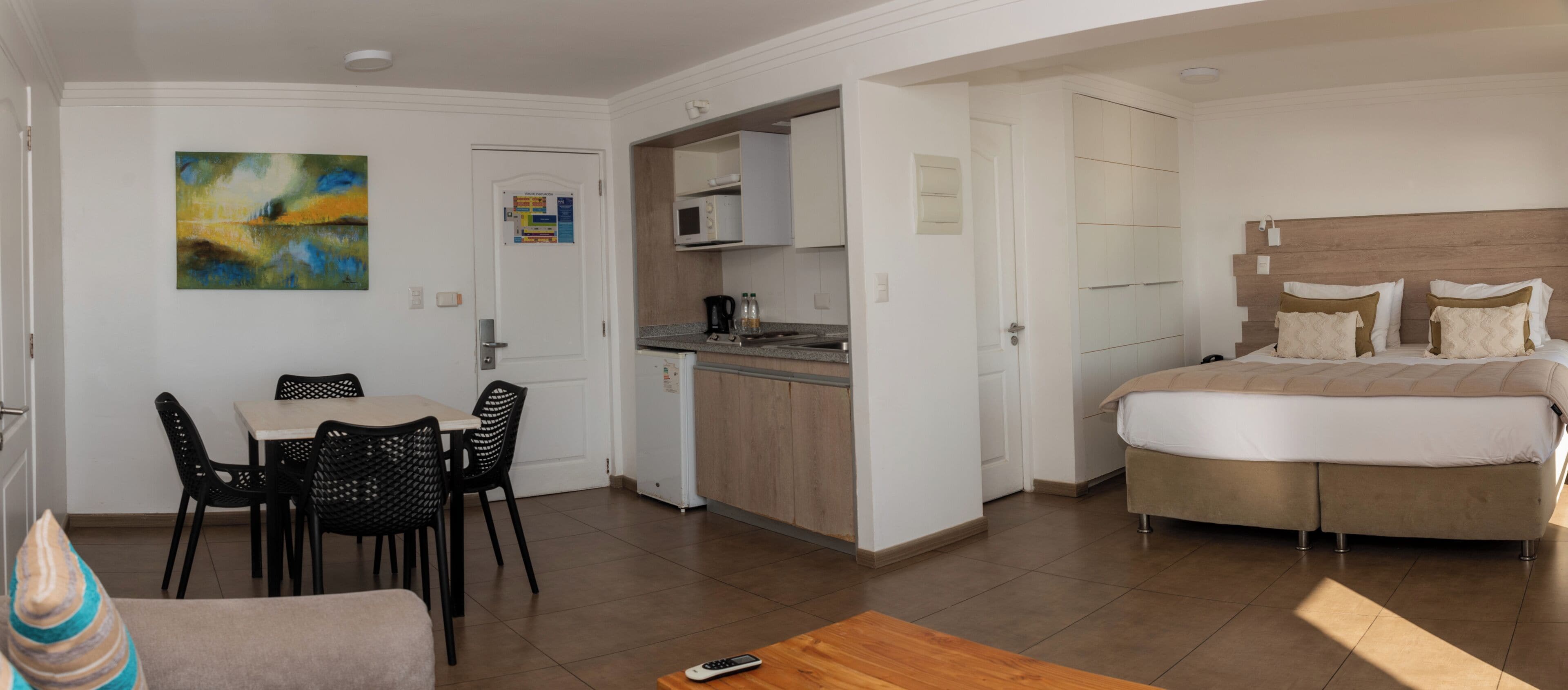Una habitación de hotel con cama, un área de comedor con sillas, una cocina y un cuadro en la pared.