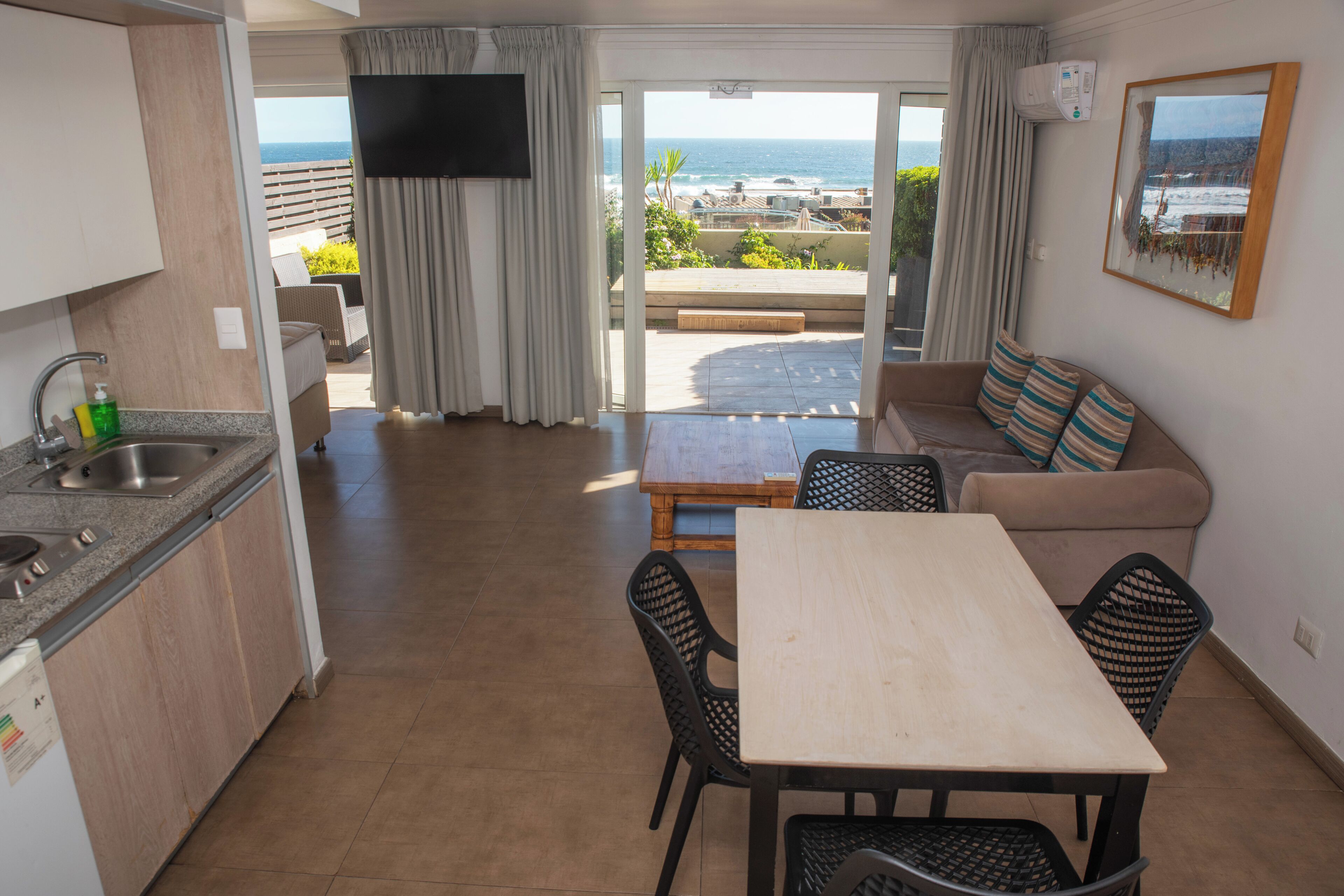 A modern kitchen with a built-in dishwasher, a dining area with a table and chairs, a sofa with striped cushions, and a large window offering a view of the ocean.