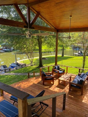 Une terrasse extérieure couverte, aménagée avec du mobilier en bois, offrant une vue sur un lac et sur des bateaux amarrés à proximité.