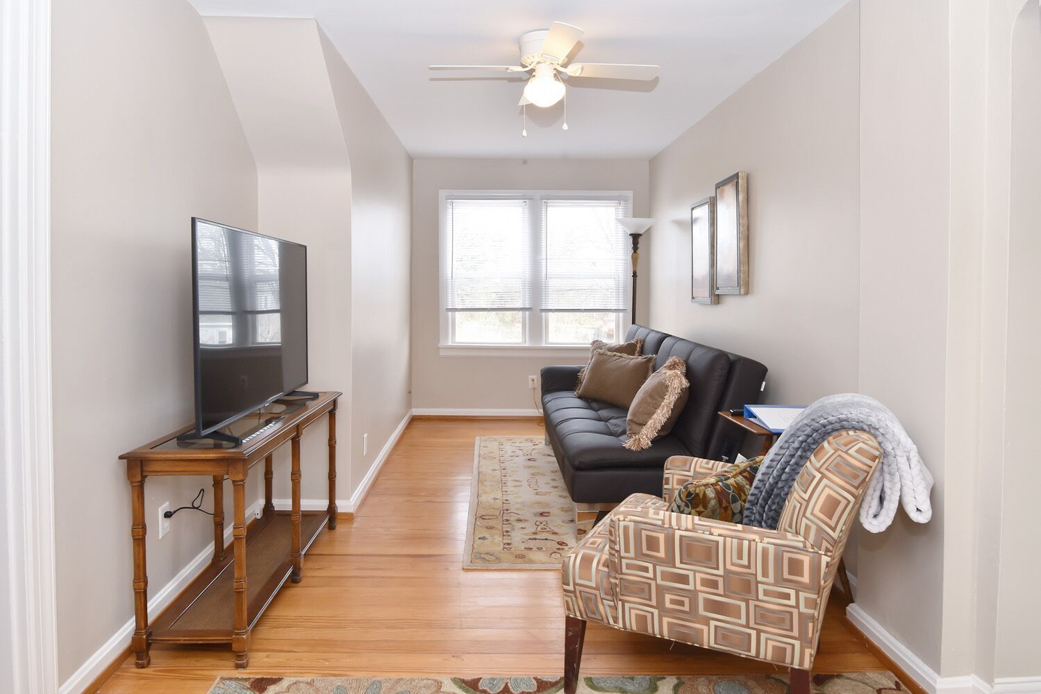A living room with a TV on a wooden stand, a sofa, a patterned armchair, and a ceiling fan.