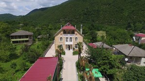 A large, two-story building with a central entrance and a red roof, surrounded by lush greenery and hills in the background. - Hotel Canyon (Khoni)
