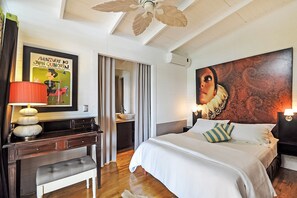 A hotel room with a bed, a desk with a lamp, a large painting, and a ceiling fan.