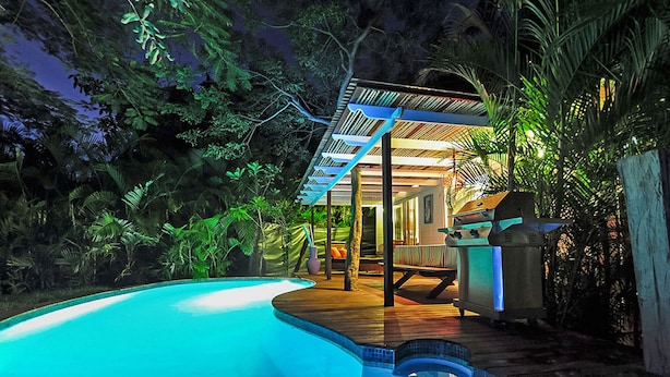 Una piscina con cubierta de madera, rodeada de abundante vegetación y un espacio de estar techado.