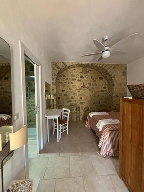 Une pièce avec un mur en pierre, un ventilateur de plafond, une table avec des chaises et deux lits avec une literie à carreaux.