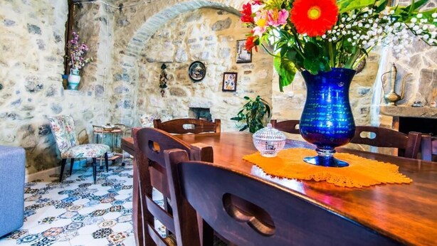 Un comedor rústico con paredes de piedra, una mesa de madera, sillas y un jarrón azul con flores.