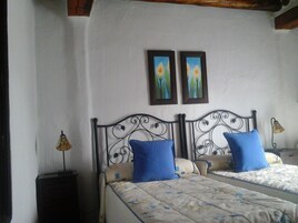 Una cama doble con una colcha estampada y dos almohadas azules, una mesita de noche con una lámpara, y dos cuadros enmarcados en la pared.