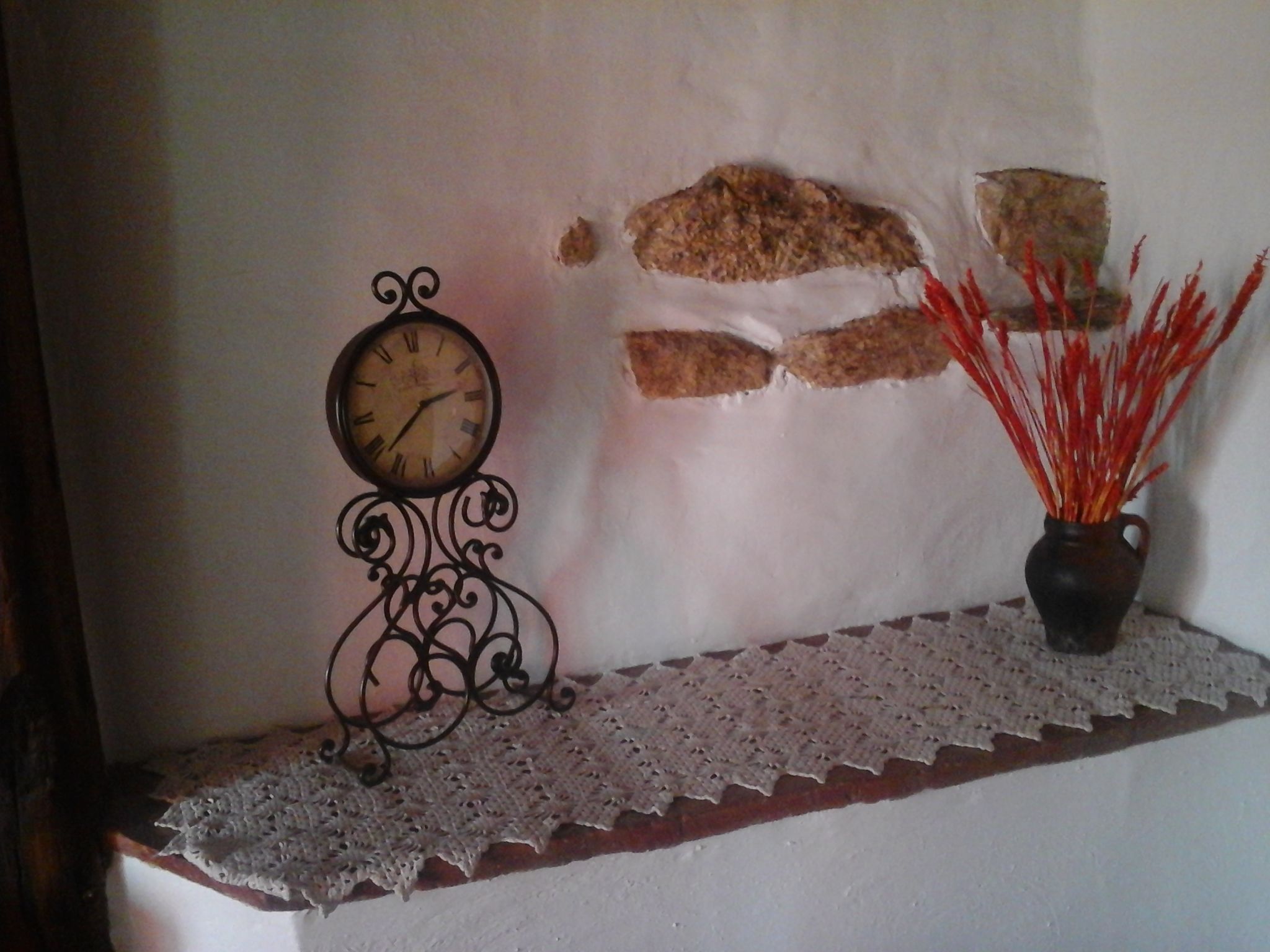 Un reloj de pared, un florero con detalles decorativos rojos y un encaje sobre una repisa.