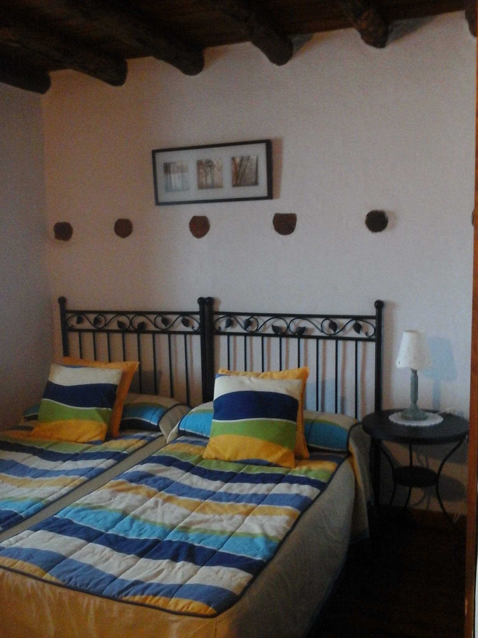 Una habitación con dos camas, cada una con ropa de cama y almohadas a rayas, una mesita con una lámpara y un cuadro en la pared.