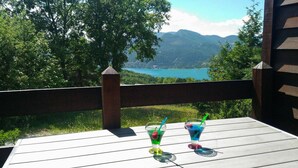 Une terrasse en bois avec deux boissons posées sur une table, donnant sur un lac et des montagnes.