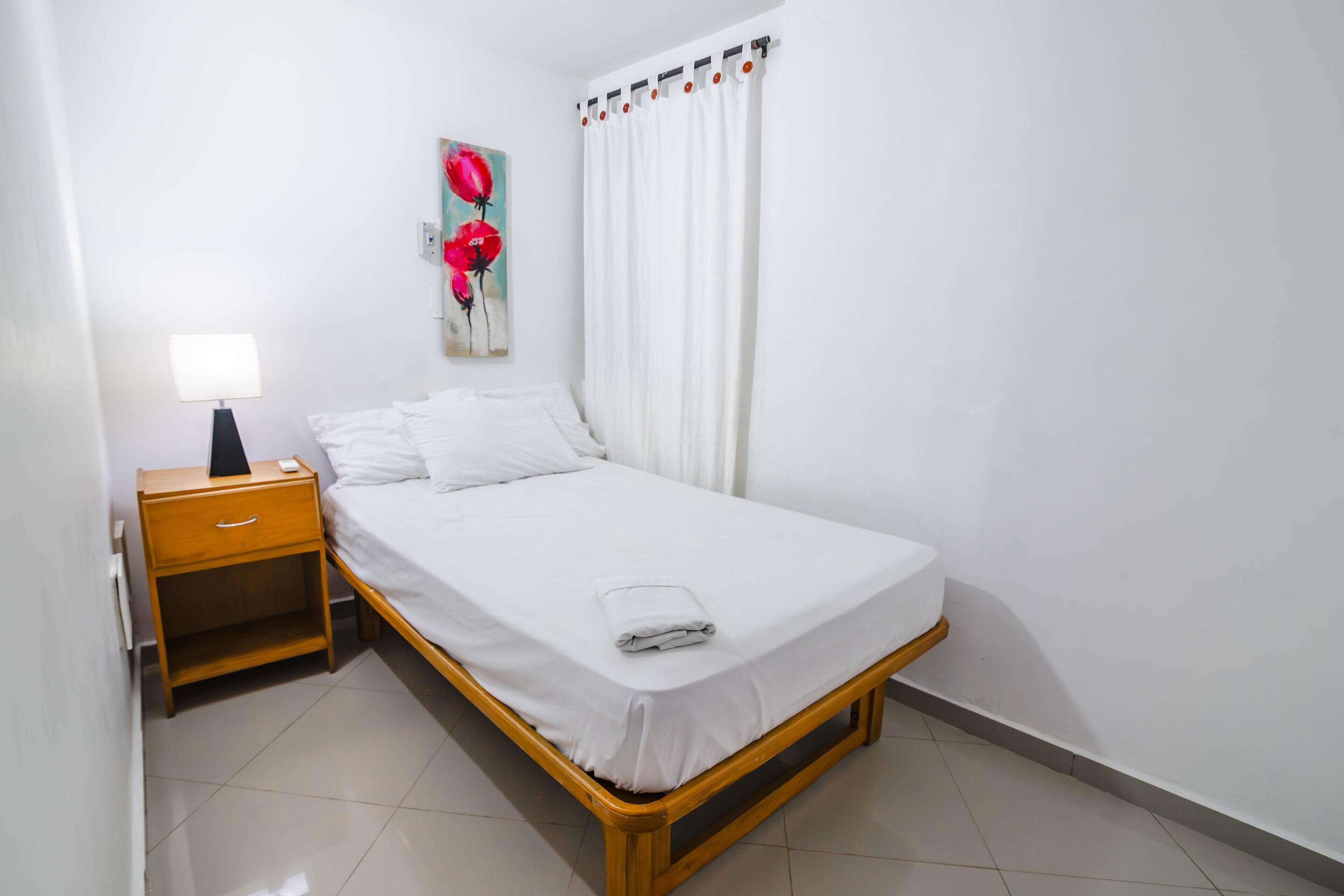 A neatly made bed with white linens, a wooden nightstand with a lamp, and a painting of red flowers on the wall.