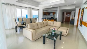 A modern living room with a sectional sofa, a glass coffee table, and a dining area with chairs and a small kitchen in the background.