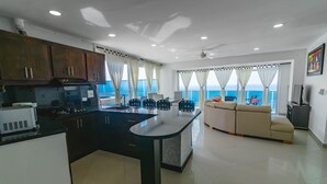 A modern kitchen with a central island, a dining area with a sofa, and large windows with a view of the sea.