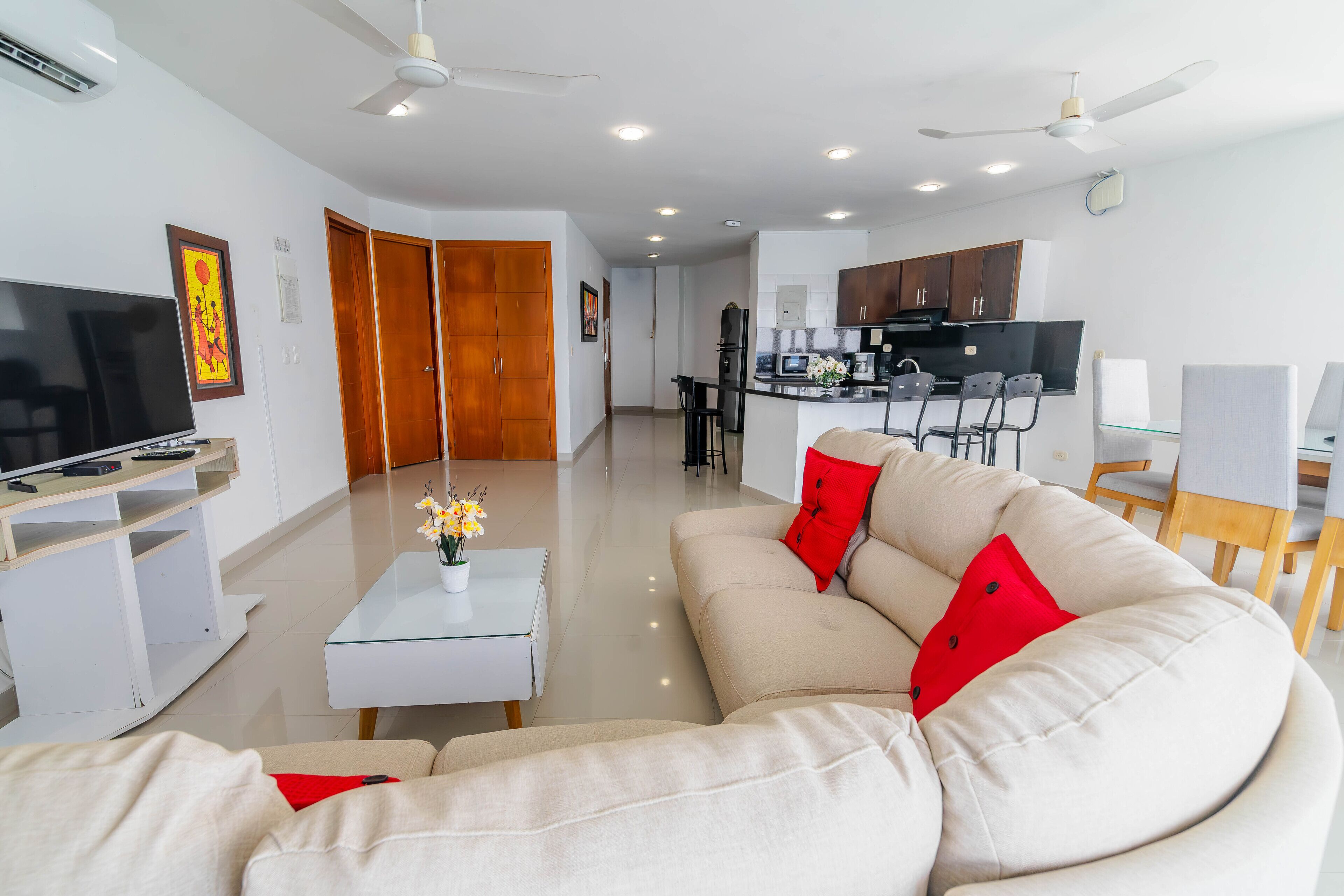 A modern living room with a sofa, a coffee table, and a TV. There is a kitchen area with cabinets and a dining table.