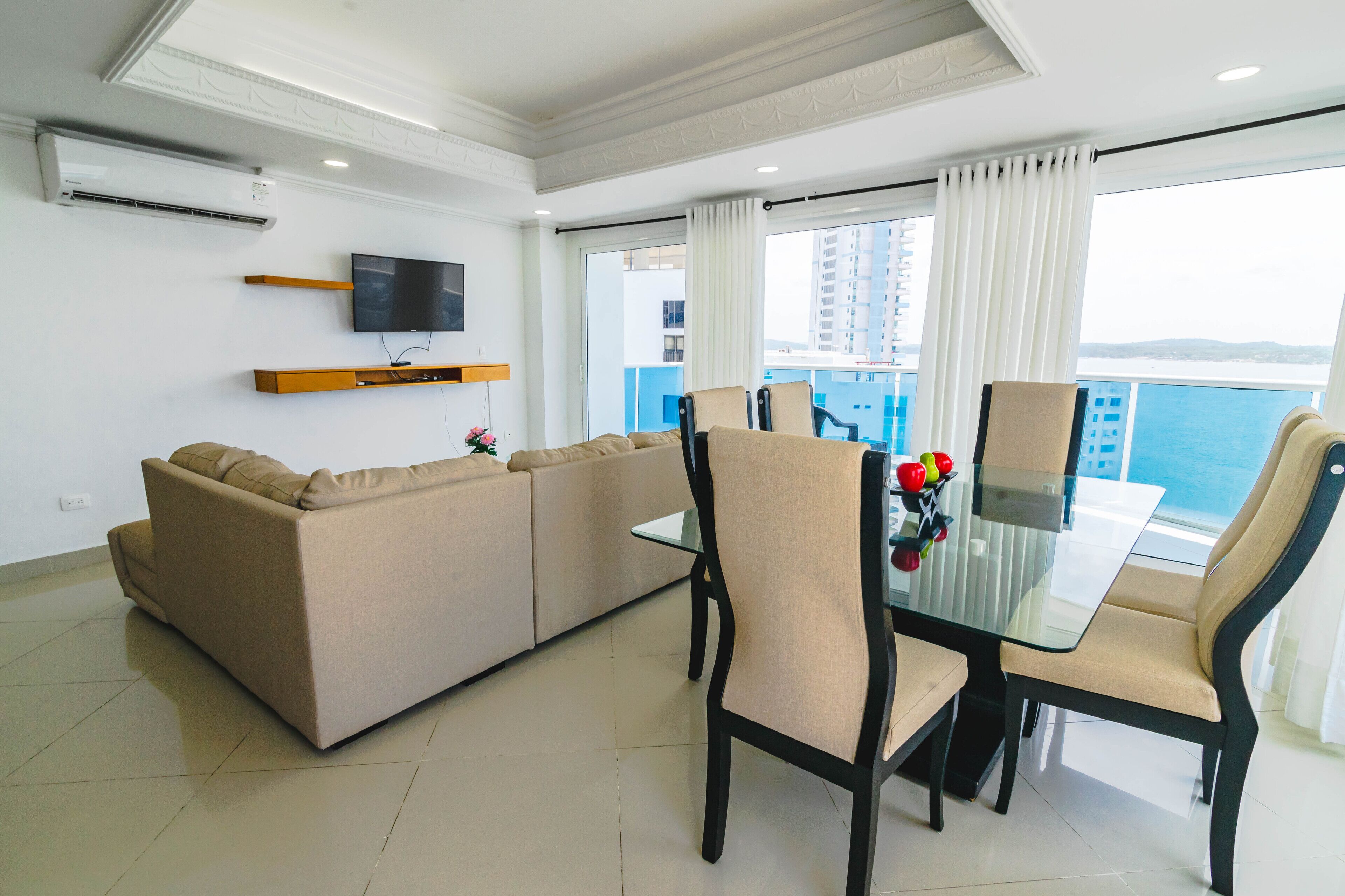 A modern living room with a sofa, a glass dining table, and chairs. There is a TV mounted on the wall and a balcony with a view of the sea.