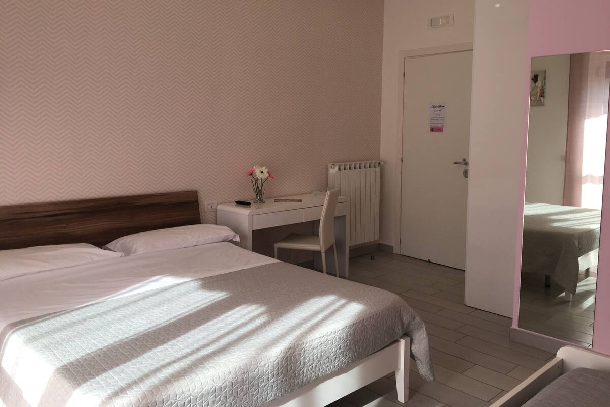 Habitación de hotel con cama, escritorio y silla. Hay un radiador debajo del escritorio y una puerta a la derecha.
