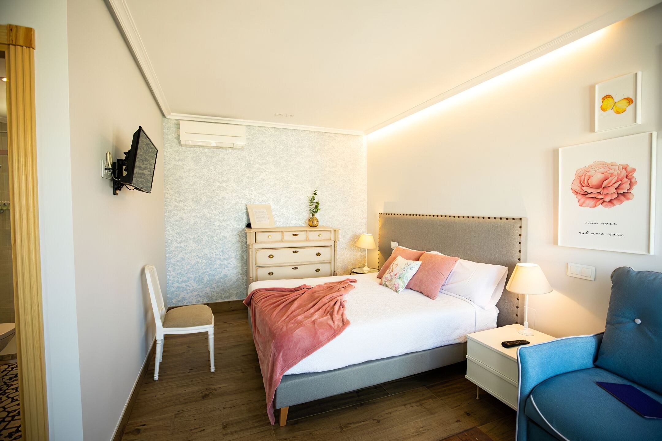 Un dormitorio con cama, mesitas de noche, una silla, cómoda y televisor montado en la pared.
