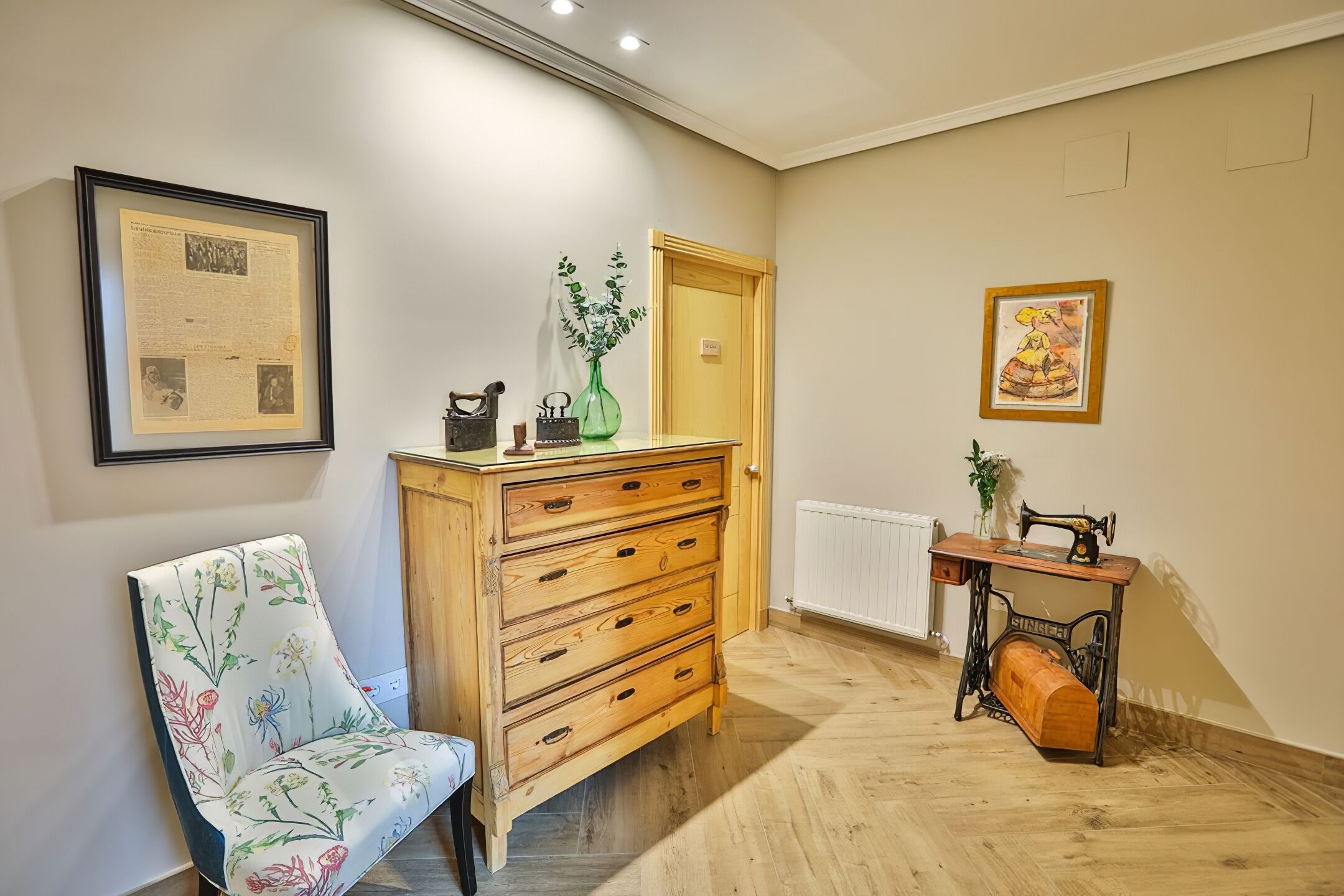 Una habitación con un aparador de madera, una silla con tapicería floral, una máquina de coser y cuadros en las paredes.