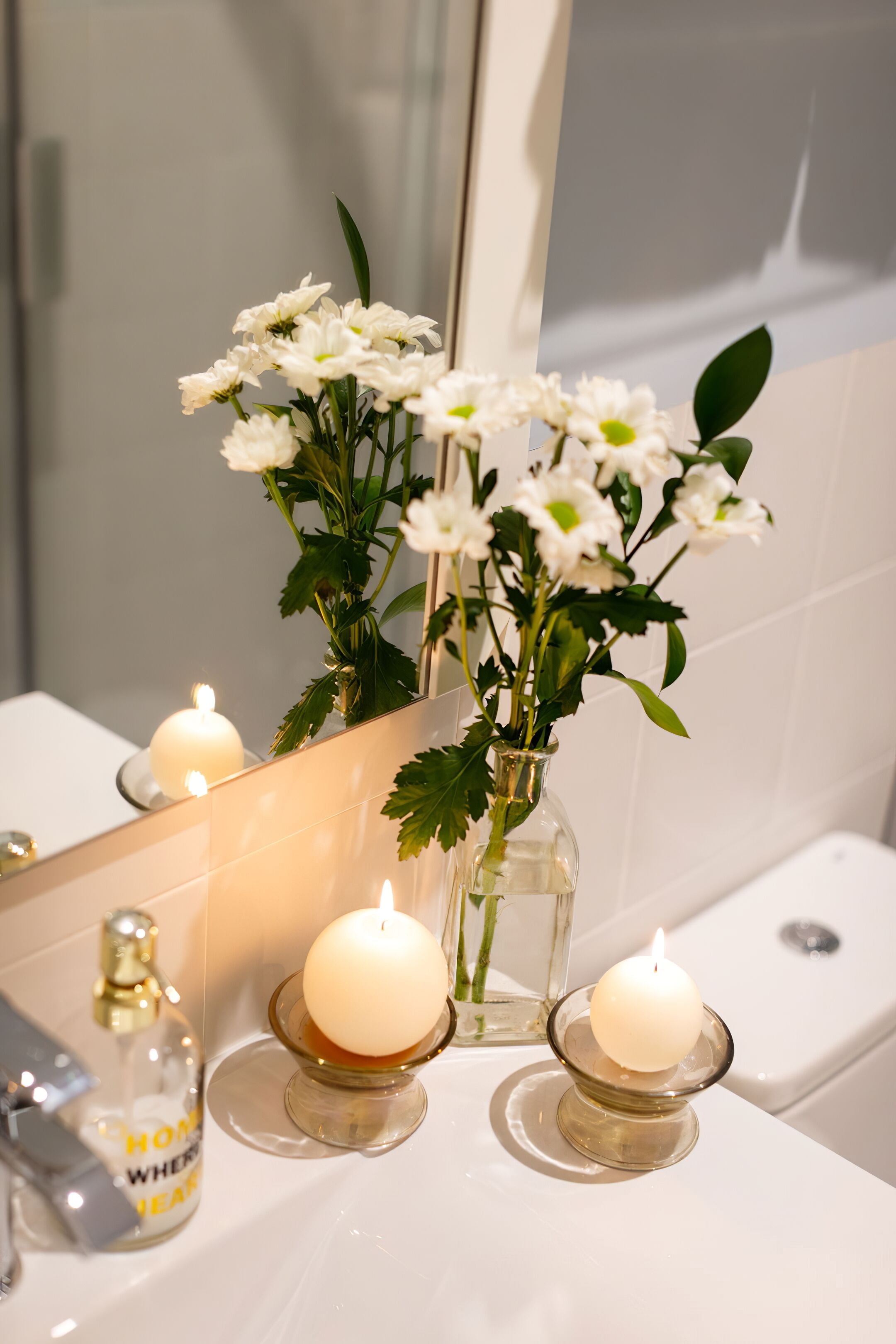 Ein Badezimmer-Waschtisch mit Spiegel, eine Vase mit weißen Blumen und zwei brennende Kerzen auf der Ablagefläche.