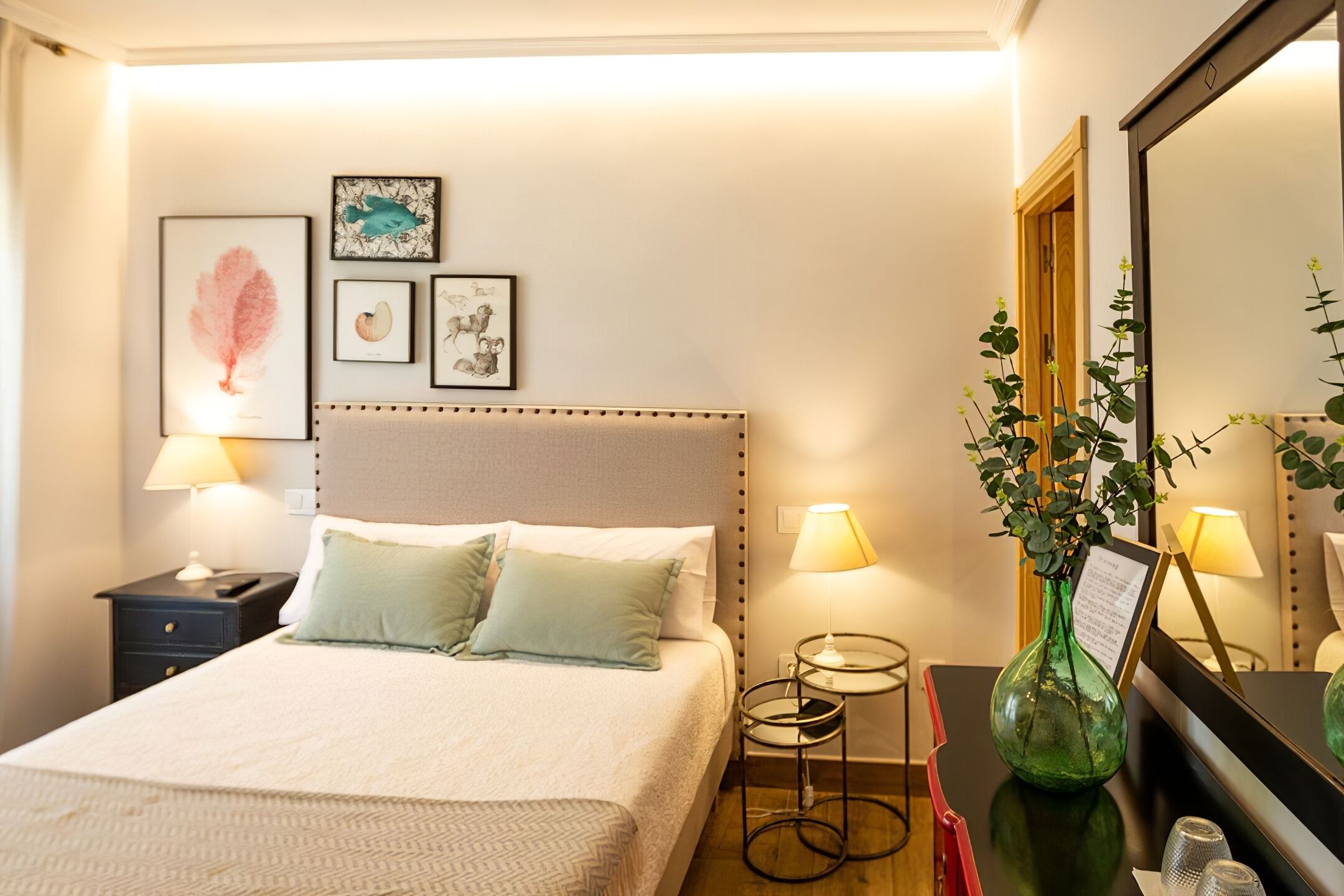 A neatly made bed with pillows, a bedside table with a lamp, a small round table with a vase of green plants, and framed artwork on the wall.