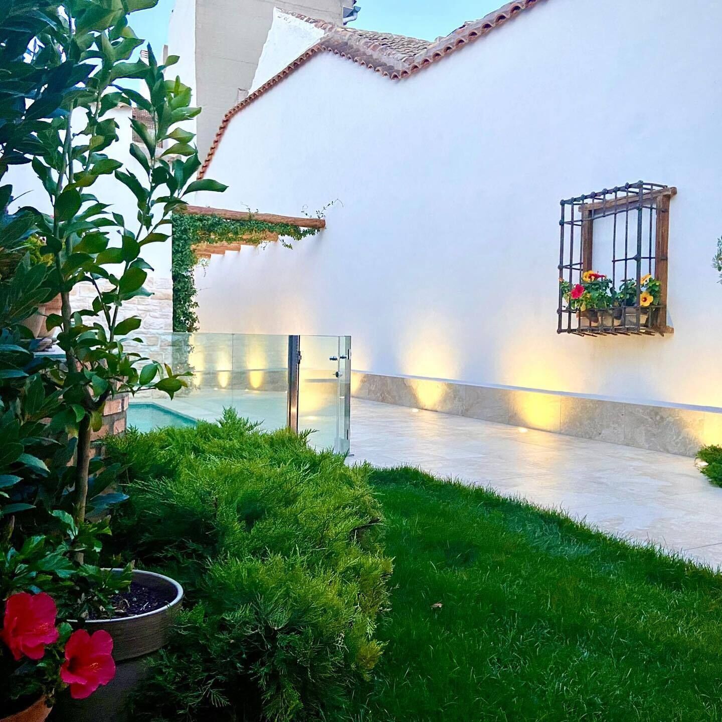 A courtyard with a white wall, a small window with flowers, and a glass door leading to a pool.