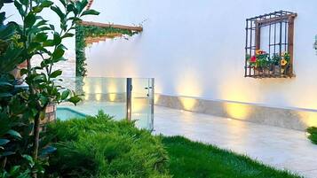 A courtyard with a white wall, a small window with flowers, and a glass door leading to a pool.
