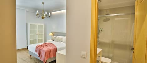 A bedroom with a bed, bedside table, and a nightstand. There is a bathroom with a shower, toilet, and sink.