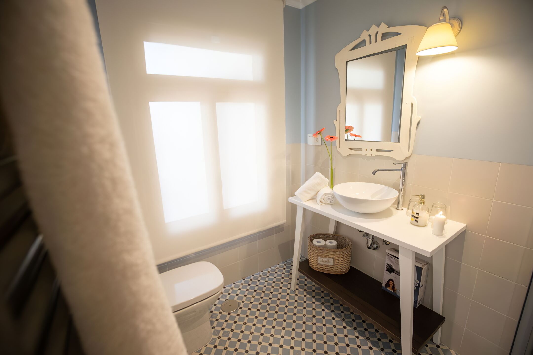Un baño con lavamanos blanco, espejo y toallero. Hay una ventana con cortinas, suelo a cuadros y una cesta con artículos de aseo.