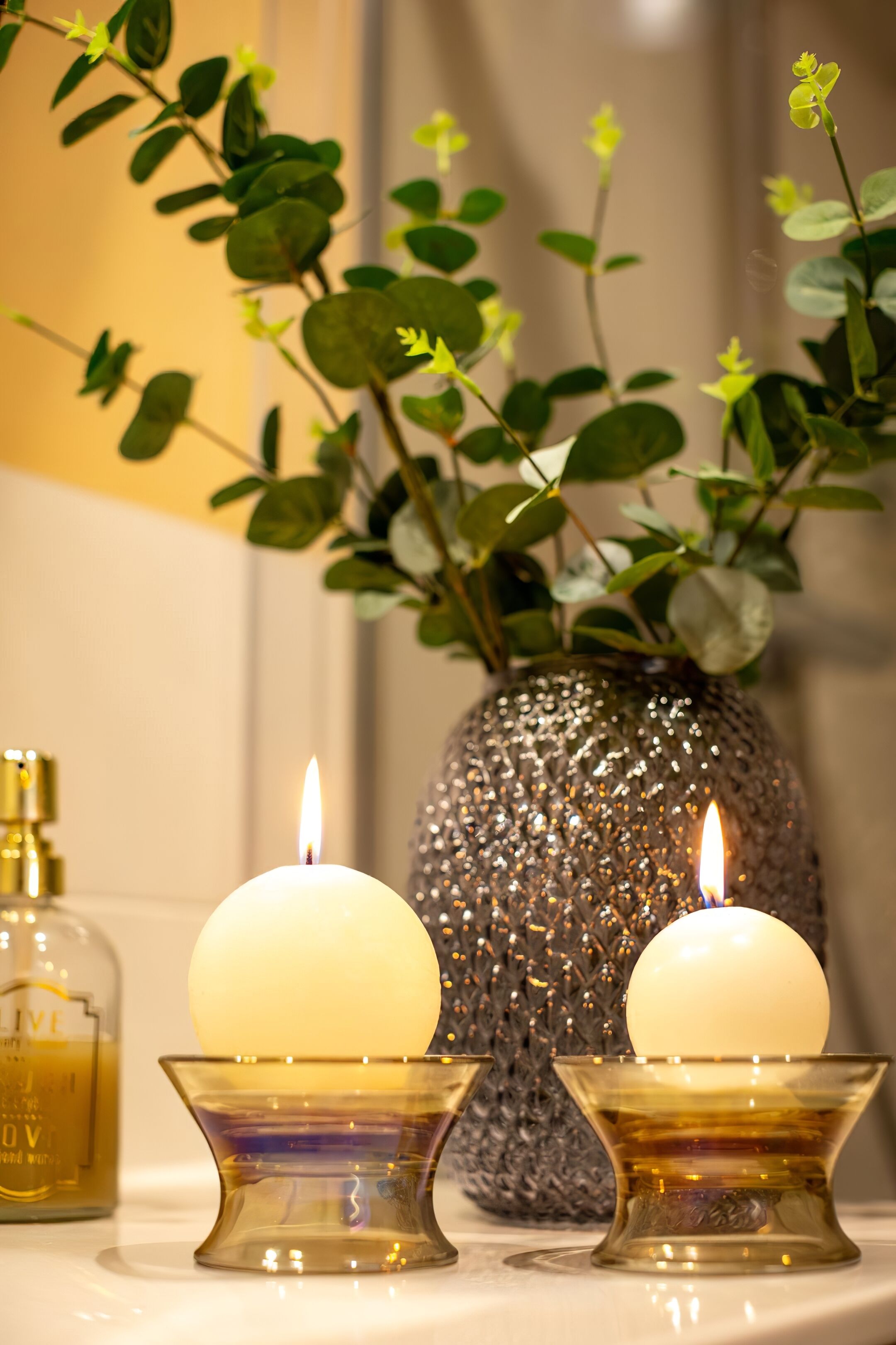 Two lit candles on glass stands, a decorative vase with green foliage, and a bottle of olive oil.