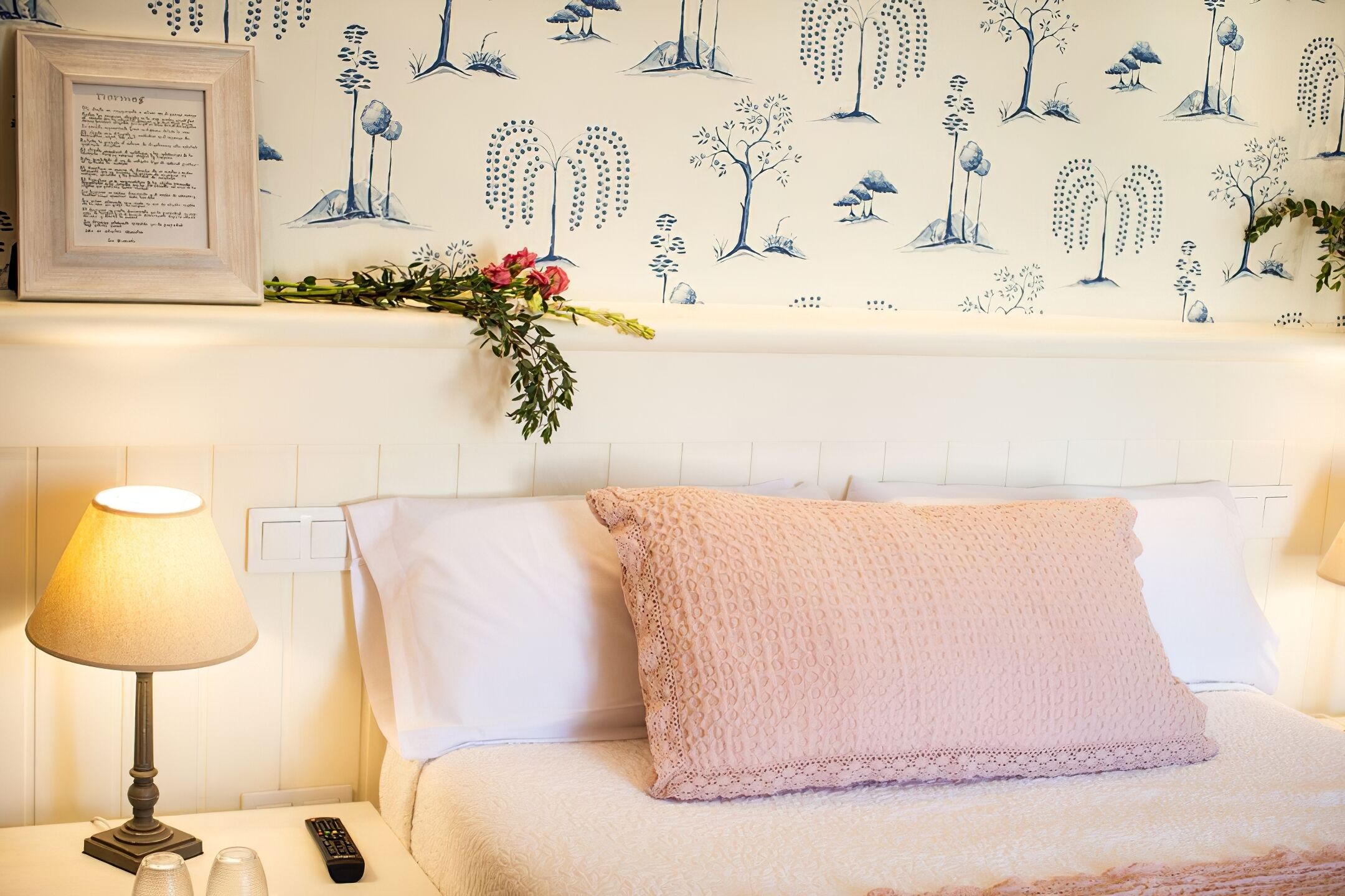 A neatly made bed with a pink pillow, a bedside lamp, a remote control, and a framed piece of paper on the nightstand.