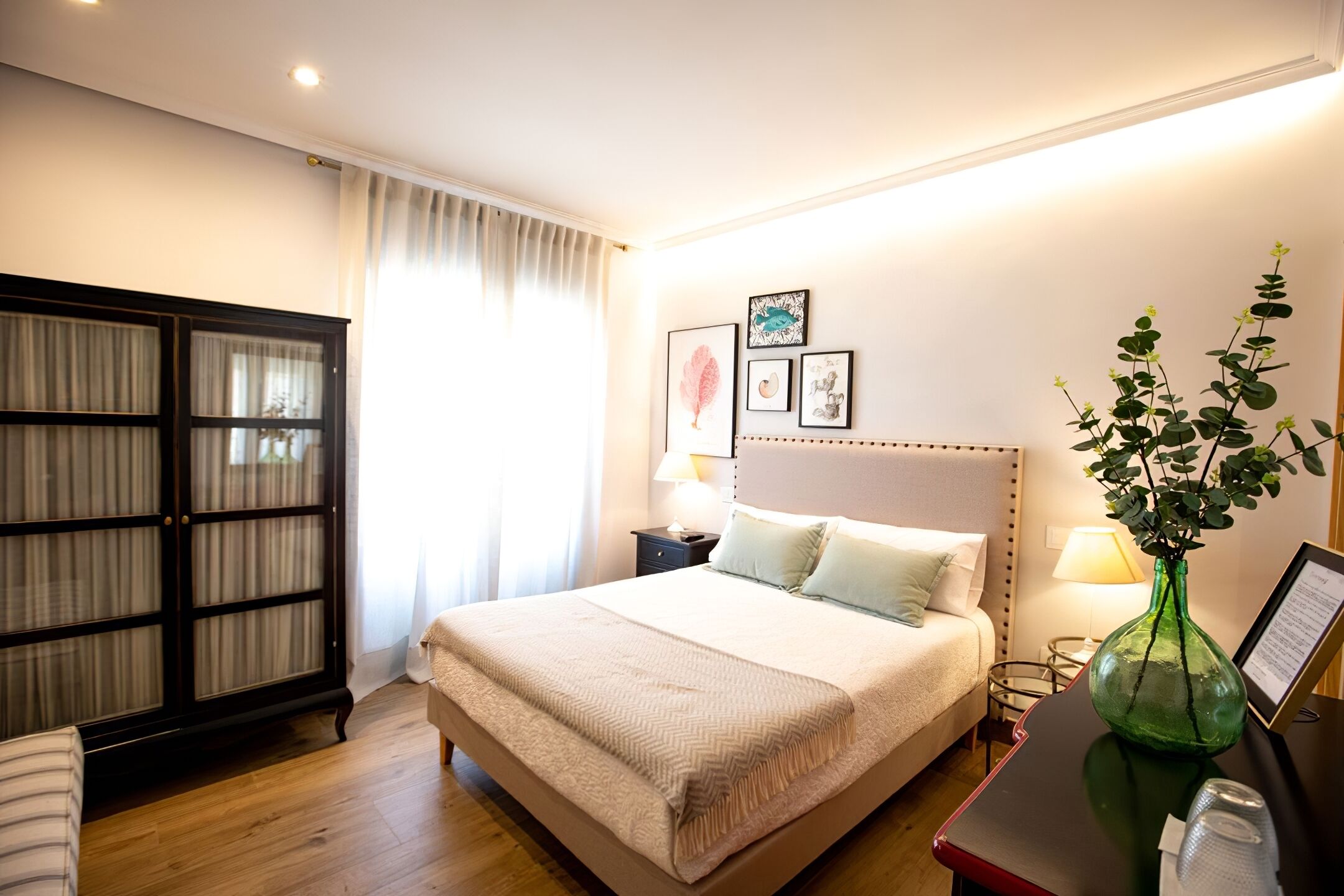 A neatly arranged hotel room with a large bed, a bedside table with a lamp, a green vase with a plant, and a wardrobe with glass doors.