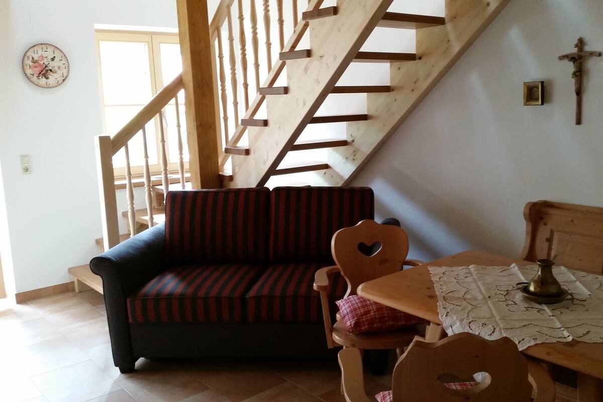 A cozy living room with a wooden staircase, a striped sofa, a wooden table with a lace tablecloth, and a cross on the wall.
