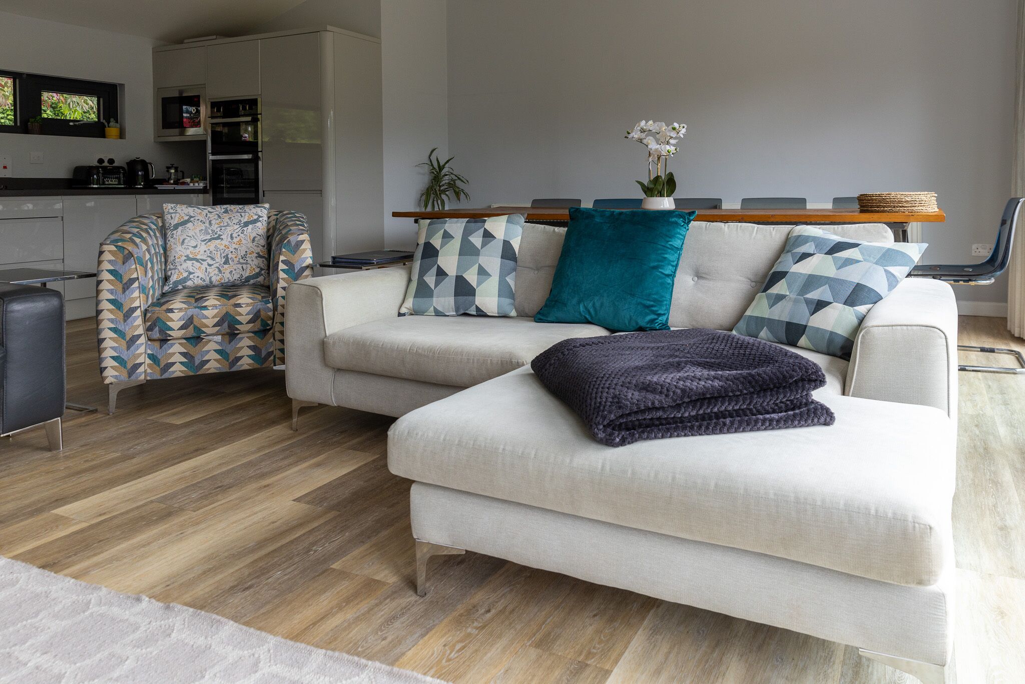 A modern living room with a sofa, armchair, and a coffee table. There is a kitchen area in the background with appliances and a dining table.