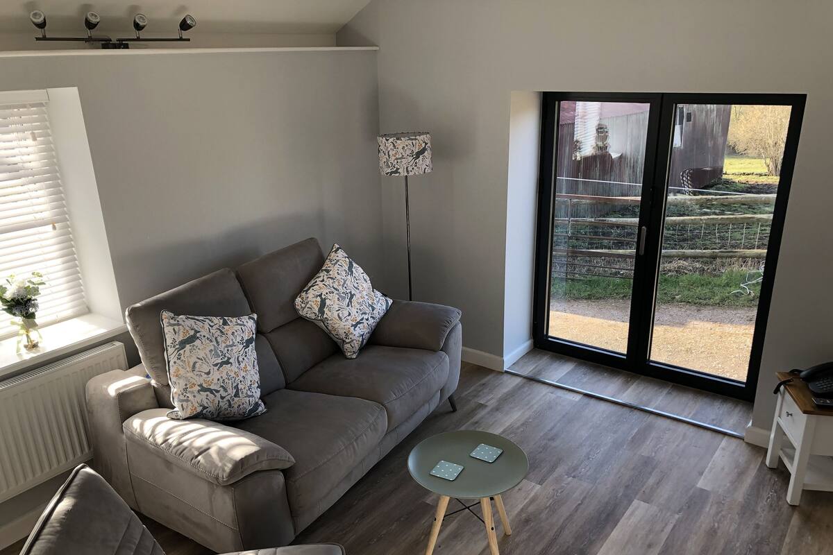 A modern living room with a grey sofa, patterned cushions, a small round table, and a view through a sliding glass door.