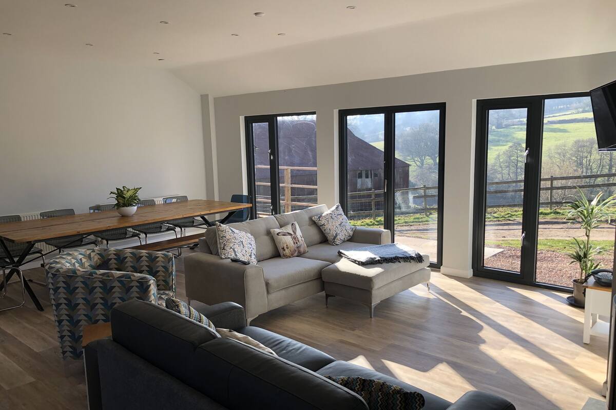 A spacious living area with a large wooden table, multiple sofas, and a view of a rural landscape through large windows.