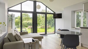 A modern living room with a sofa, a dining table, and chairs. There is a large window with a view of greenery outside. - Welsh Marches at Upper Glyn Farm (Chepstow)
