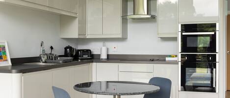 A compact kitchen with white cabinets, a built-in oven, a coffee maker, and a small dining table with chairs.