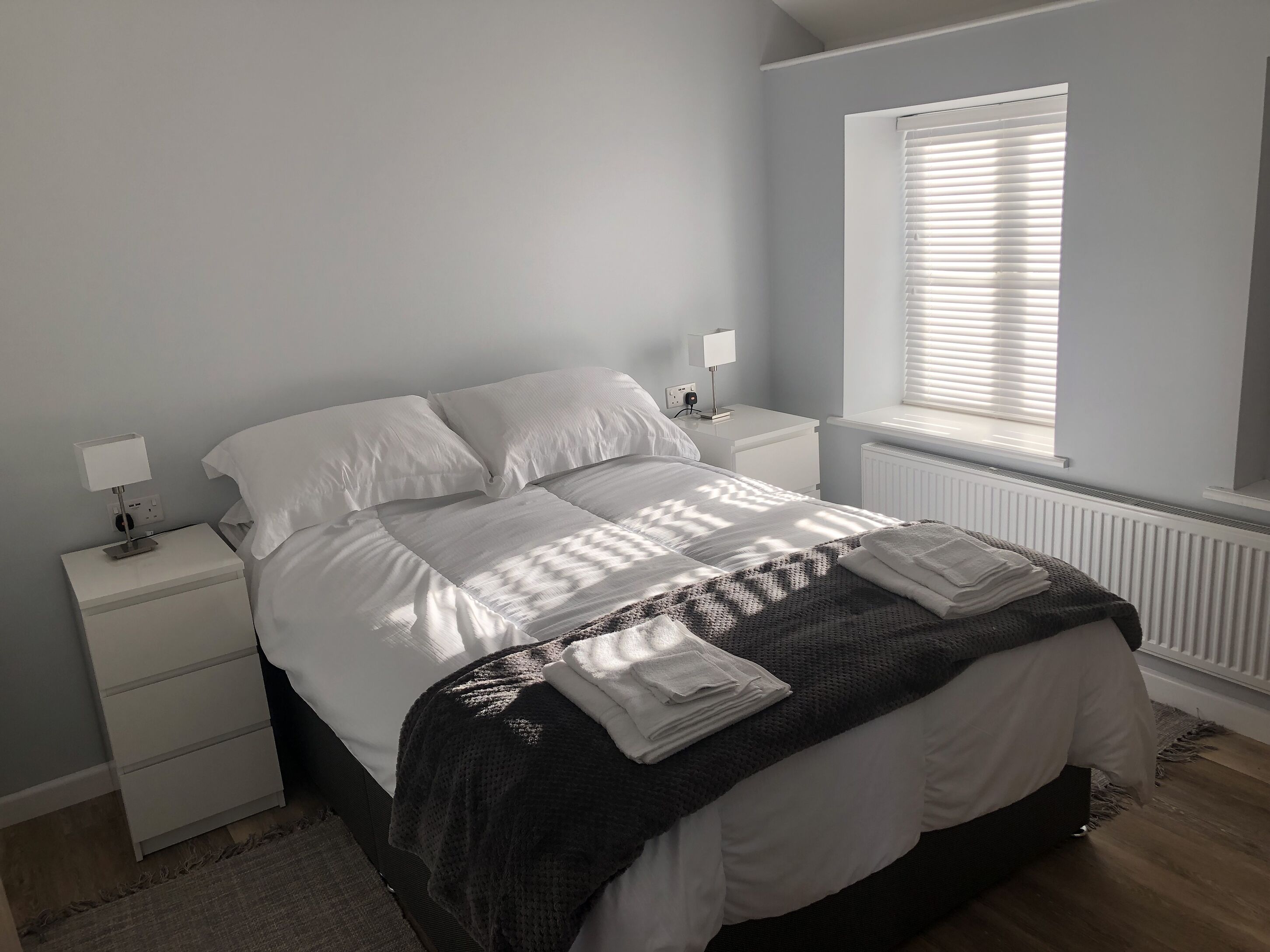 A neatly made bed with white linens, a dark grey blanket, and folded towels on a wooden floor.
