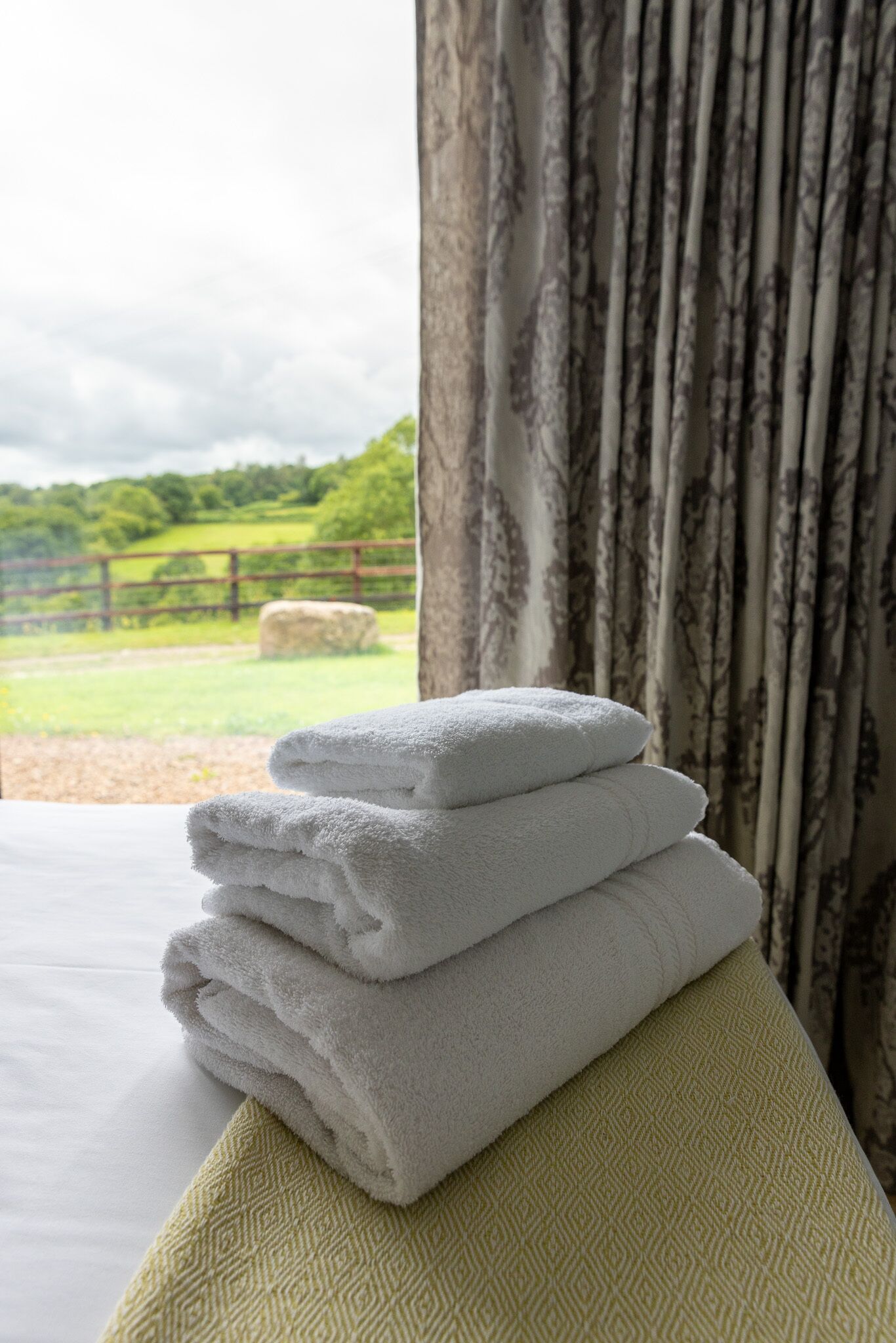 A stack of white towels on a bed with a view of a green landscape through a window with curtains.