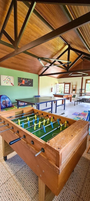 A foosball table in a room with a high ceiling and wooden beams, a pool table, and a colorful mural on the wall. - Esquilo Hotel (Camanducaia)