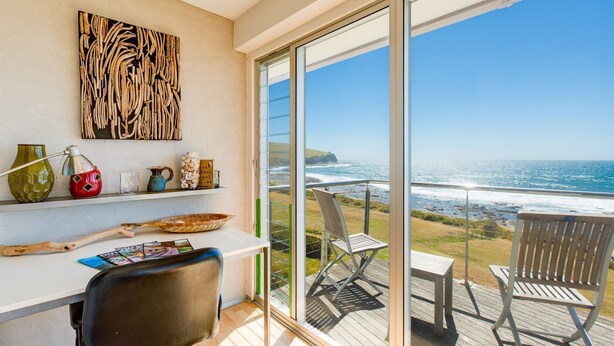 A balcony with a view of the ocean, a wooden table, a chair, and a shelf with decorative items.