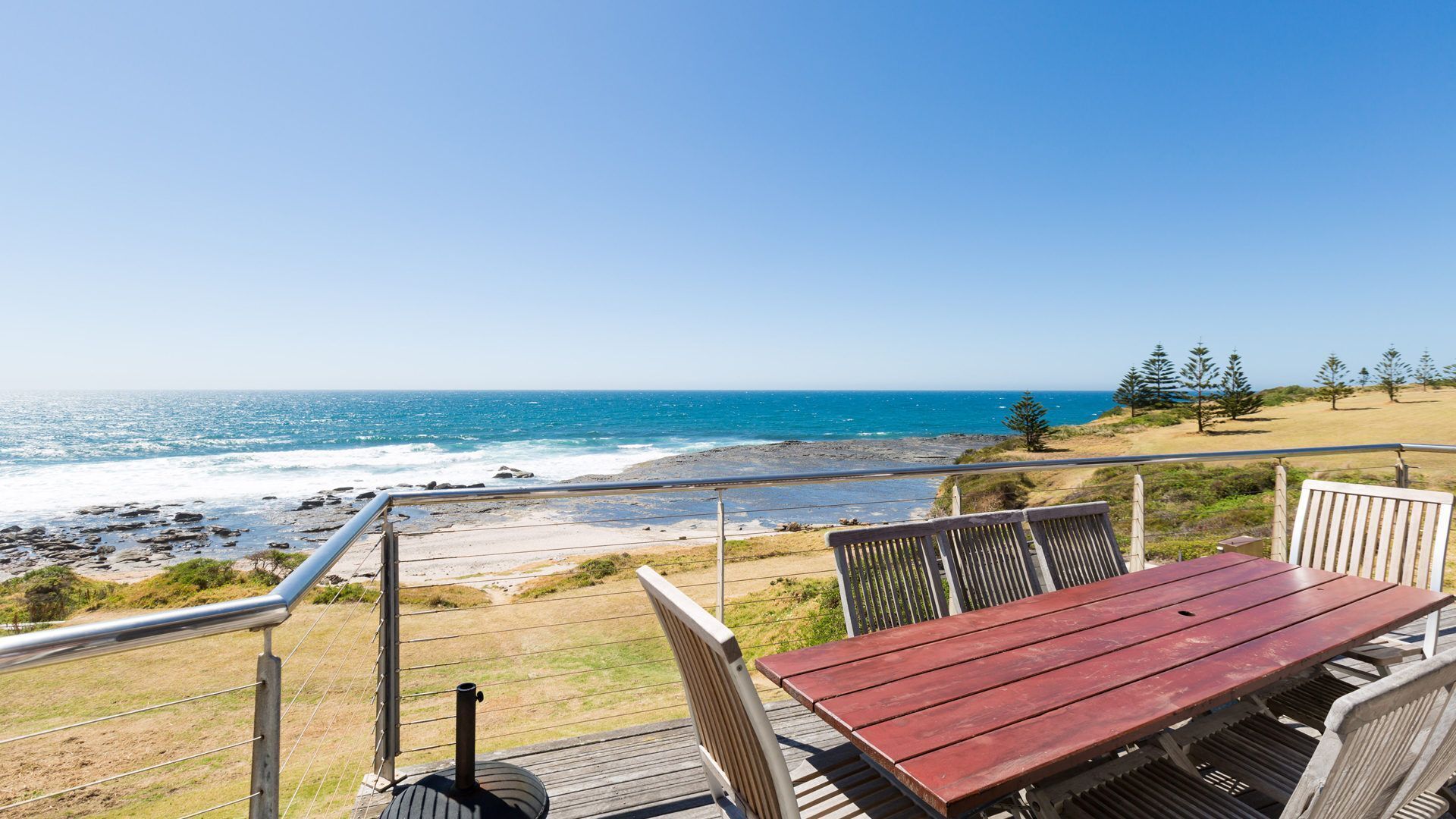 A wooden deck with a red table and chairs overlooking a rocky beach and ocean.