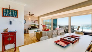 A modern living room with a large TV, a dining area with a table and chairs, a red cabinet, and a balcony with a view of the ocean.