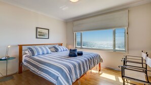 A bedroom with a bed, a bedside table, a lamp, a window with a view of the sea, and a picture on the wall.