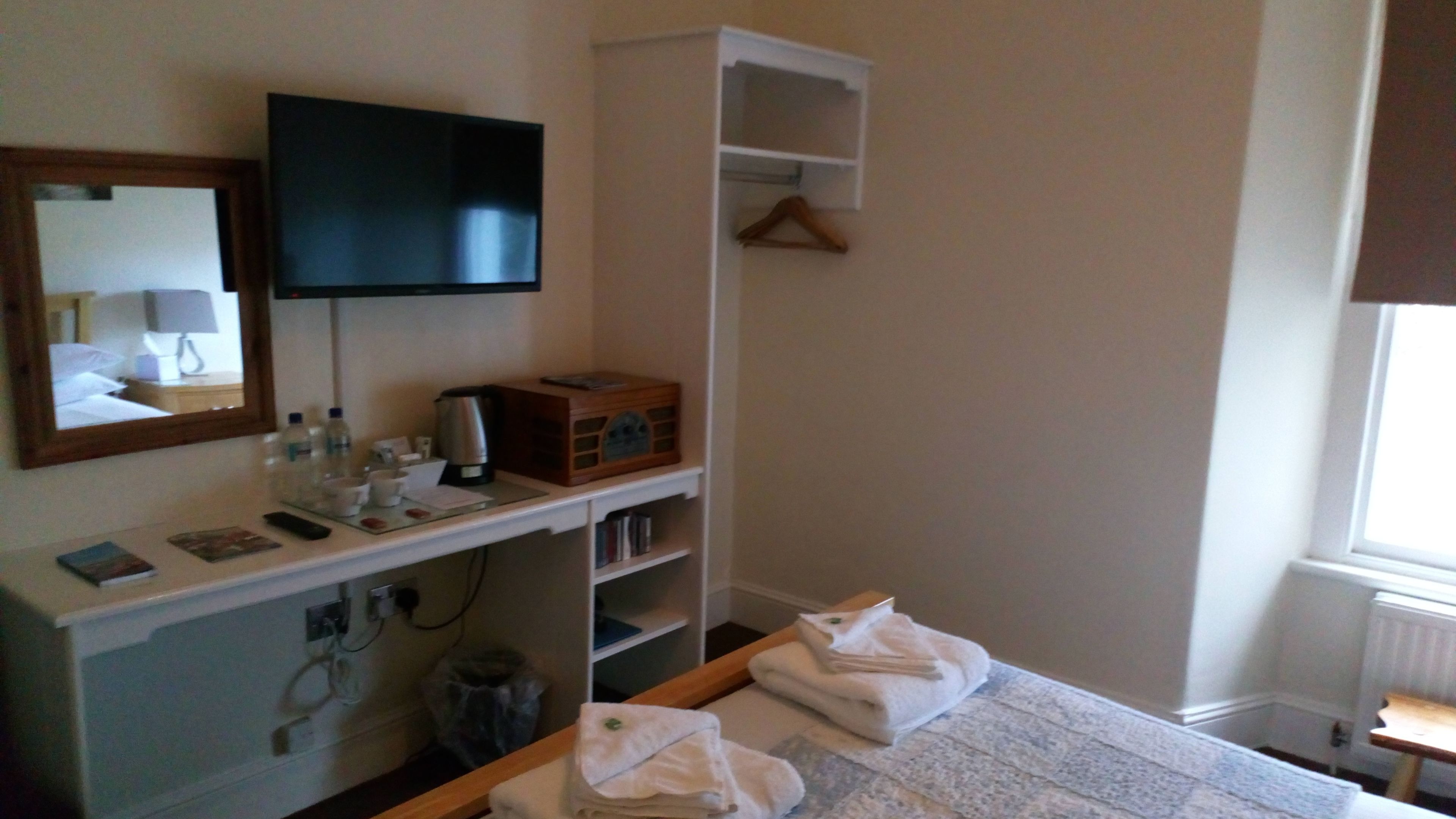 Habitación de hotel con una cama, un televisor montado en la pared, una mesita de noche con una tetera, una radio y un espejo.