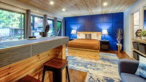 A modern bedroom with a wooden ceiling, a large bed, two bedside lamps, a desk with a chair, and a view of the outdoors through large windows.