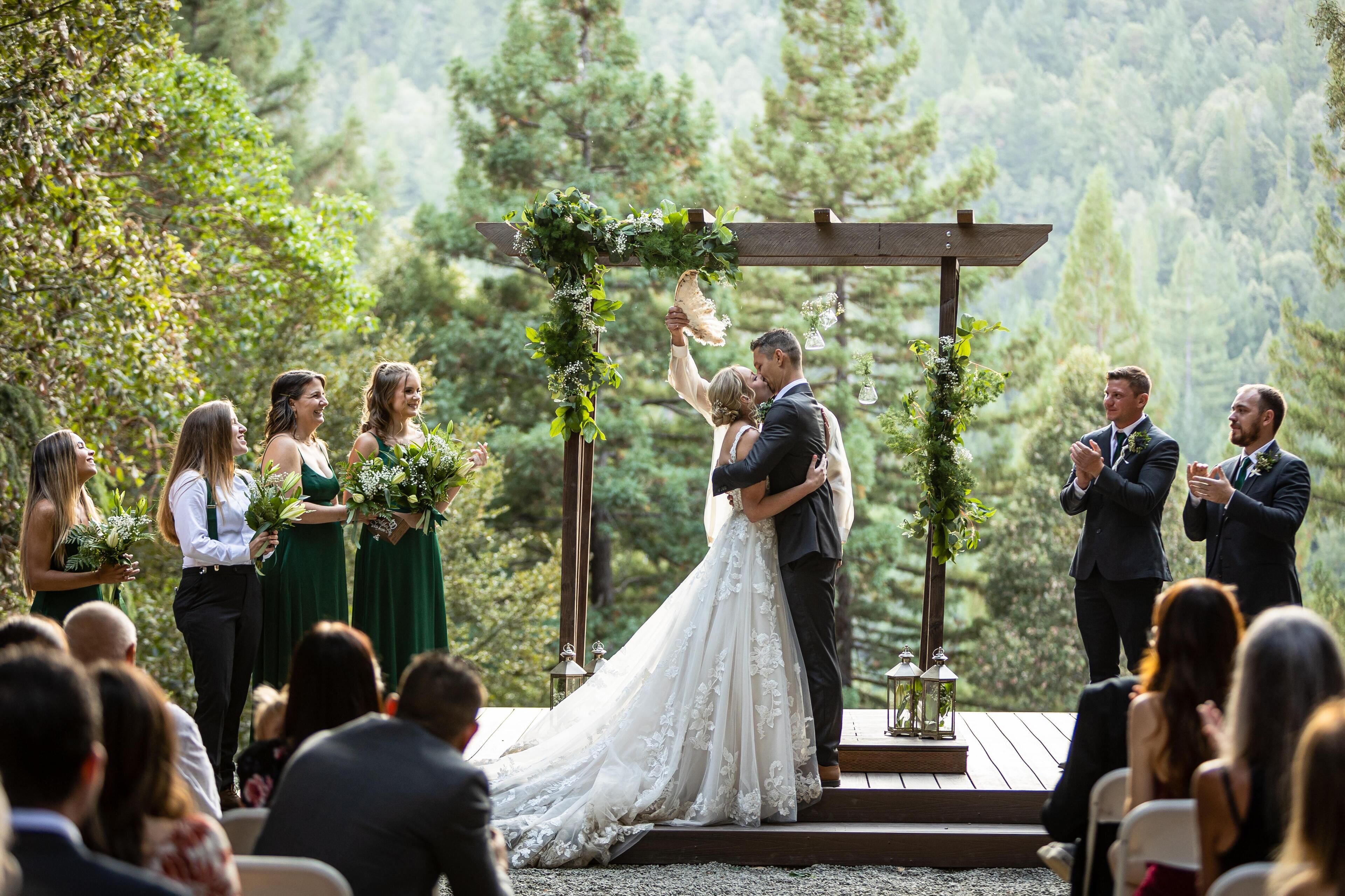 A wedding ceremony outdoors with a wooden arch and greenery.