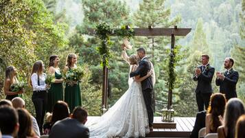 A wedding ceremony outdoors with a wooden arch and greenery.