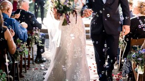 A wedding ceremony with guests seated on wooden chairs, a bride and groom walking down the aisle, and a backdrop of trees and mountains.